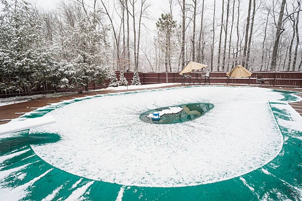 The image shows a backyard swimming pool covered with a winter pool cover and a layer of light snow. The surrounding area is snowy, and the trees in the background are bare, indicating a winter setting. A small open patch of water is visible in the middle of the pool cover, possibly due to melted snow or rainwater accumulation.